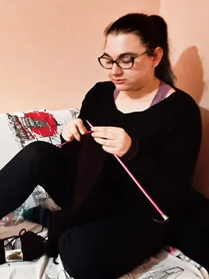 A woman sitting on a bed knitting a pair of socks.