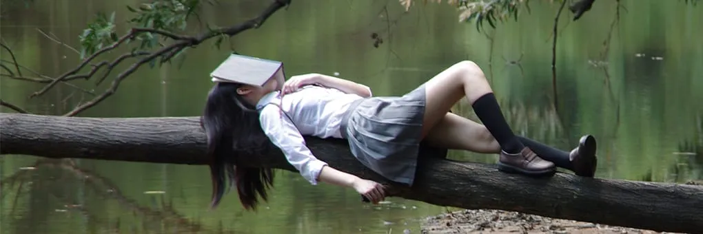 A girl laying on a tree branch reading a book.