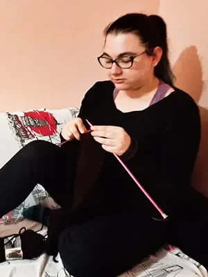 susam A woman with glasses and dark hair tied back sits cross-legged on a bed, wearing black clothes. She is focused on knitting with pink knitting needles and dark yarn. The wall behind her is a soft peach color.
