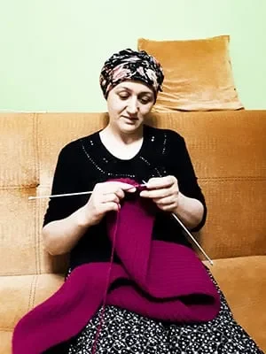 susam A woman wearing a patterned headscarf and black top sits on a tan couch, knitting a vibrant magenta garment with two knitting needles. She looks down, concentrating on her handiwork. A square, orange cushion is behind her on the couch.